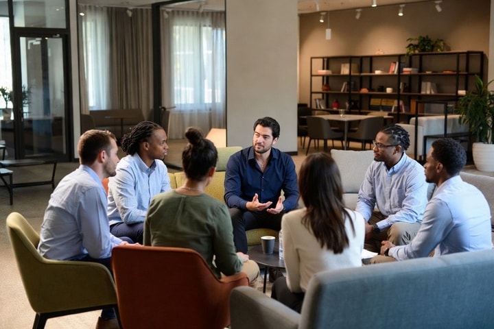 A diverse group of employees in a modern office lounge engaged in an attentive, supportive conversation — representing workplace chaplaincy and teams who feel truly seen