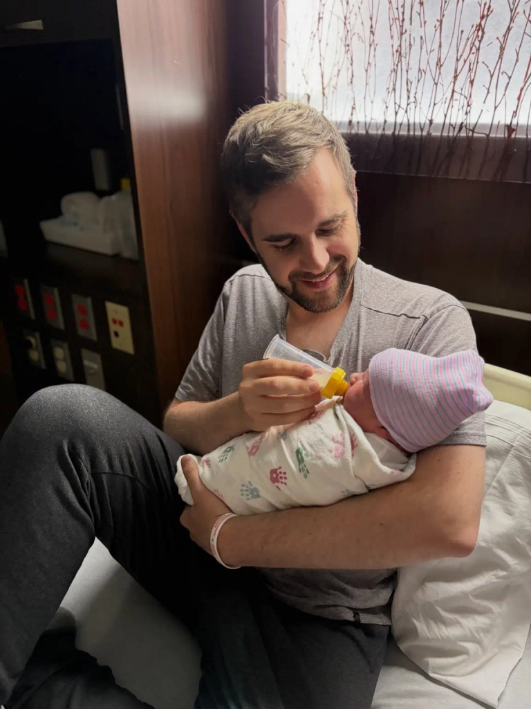A man bottle-feeding a newborn girl in a hospital room
